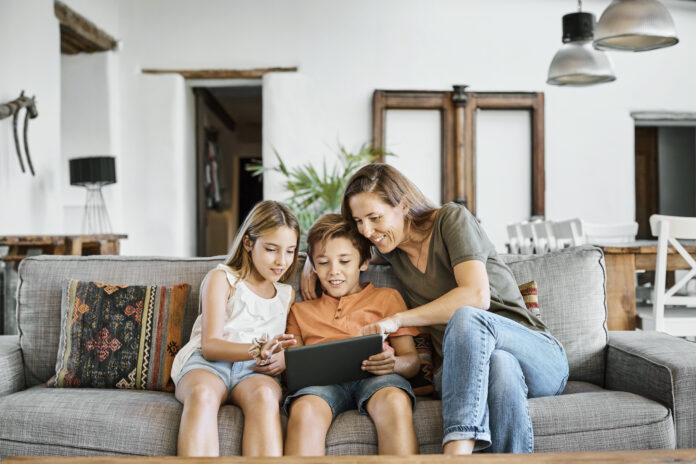 Smiling mother assisting children while showing digital tablet. Family is sitting on sofa in living room. They are in casuals at home.