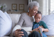 A beautiful senior woman sits on a couch with her granddaughter as they read a story together while smiling.